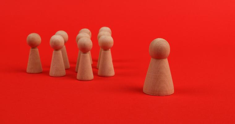 A group of wooden figurines on a red background, with one taller figure standing apart from six smaller figures.