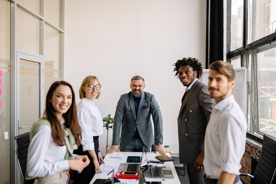 Five adults gather around a table covered with office materials in a bright meeting room.