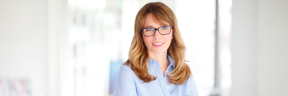 Woman wearing glasses and a light blue shirt standing with crossed arms beside a laptop in a bright office.