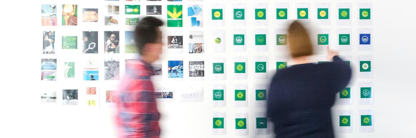 Two people interact with a wall of tennis-related images and logos, one reaching out to a logo while the other walks by.