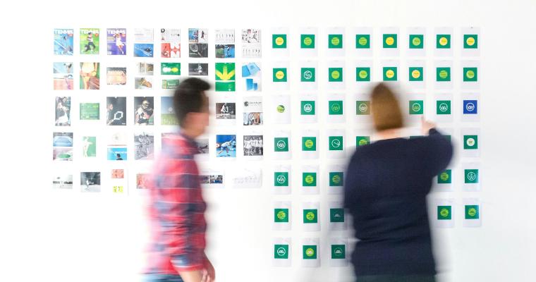 Two people interact with a wall of tennis-related images and logos, one reaching out to a logo while the other walks by.