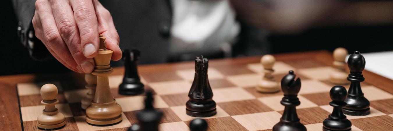 A hand moves a light-colored chess piece on a wooden chessboard surrounded by black and tan pieces.