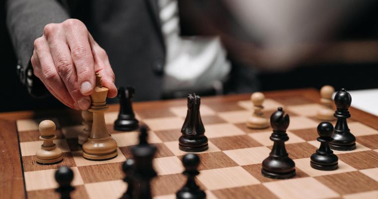 A hand moves a light-colored chess piece on a wooden chessboard surrounded by black and tan pieces.