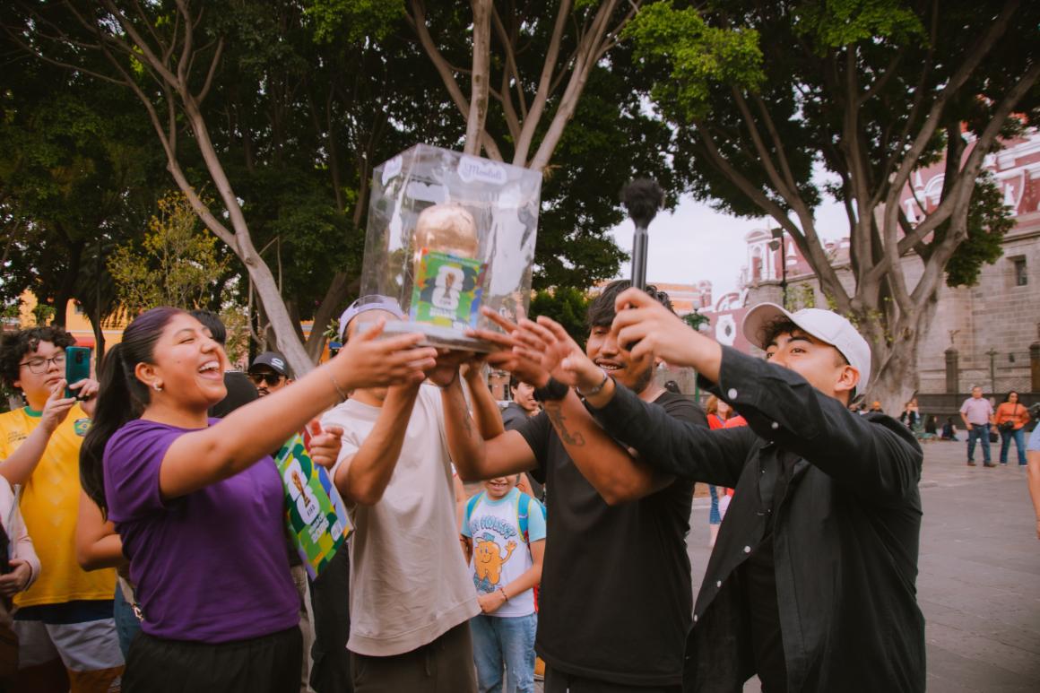 A group of young people celebrates while holding a trophy in a clear case, surrounded by trees and other spectators.