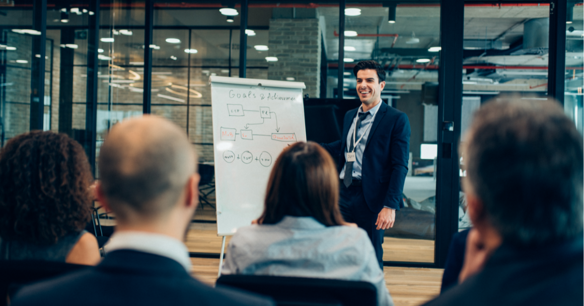 A presenter in a dark suit smiles while addressing an audience in a modern office. A whiteboard displays a flowchart titled Goals & Achievements.