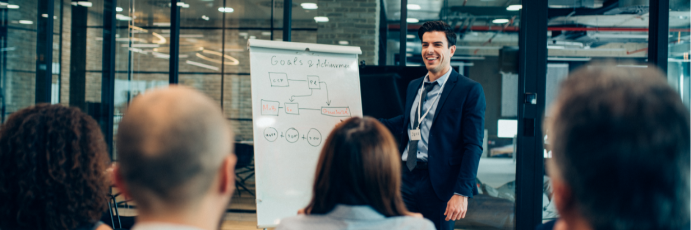 A presenter in a dark suit smiles while addressing an audience in a modern office. A whiteboard displays a flowchart titled Goals & Achievements.
