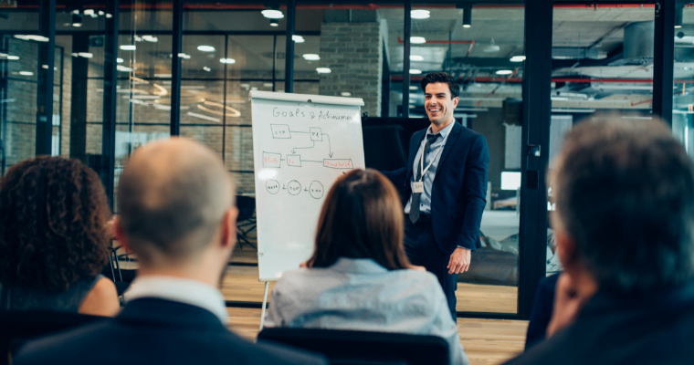 A presenter in a dark suit smiles while addressing an audience in a modern office. A whiteboard displays a flowchart titled Goals & Achievements. image link to article