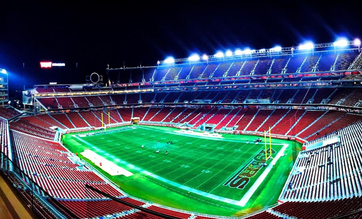 A nighttime view of an empty football stadium with illuminated green turf and red seating. Scattered figures are seen on the field.