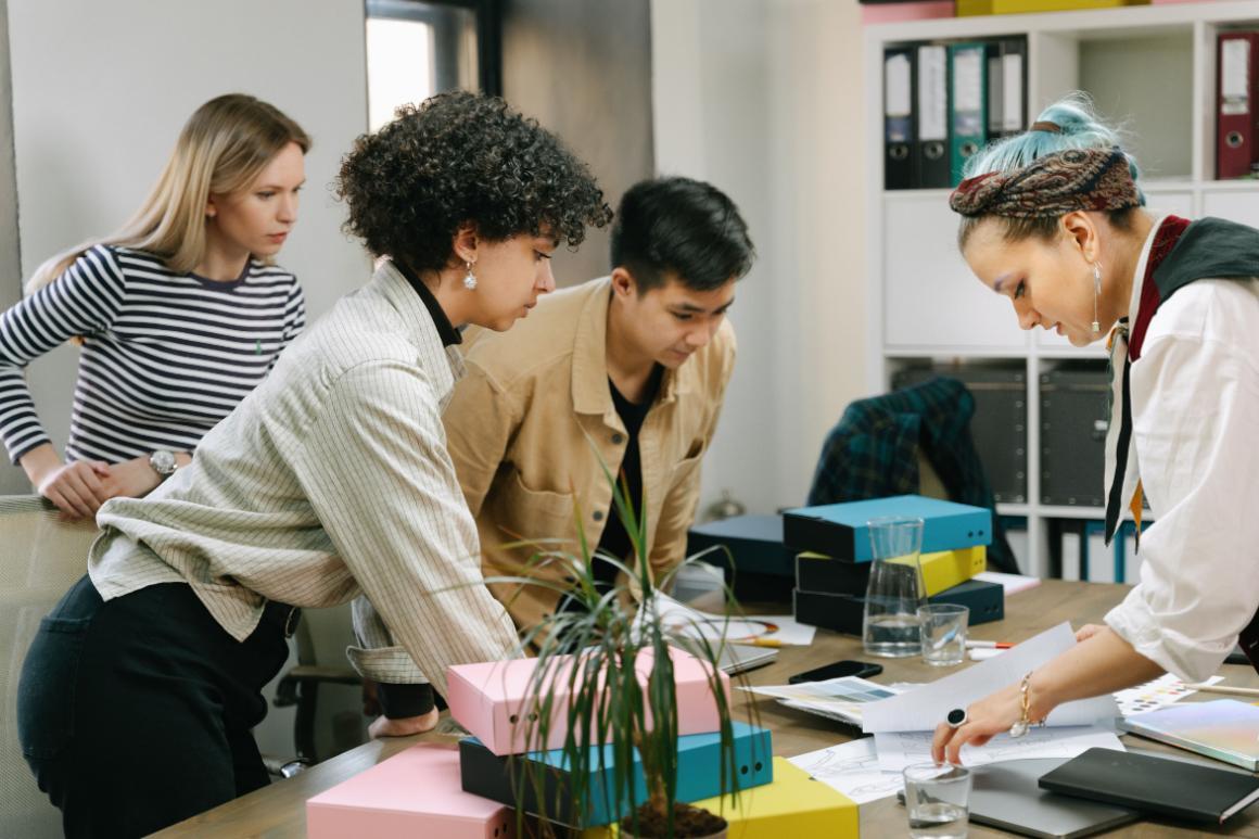 Four people collaborate around a table covered with documents, design materials, storage boxes, and a potted plant.