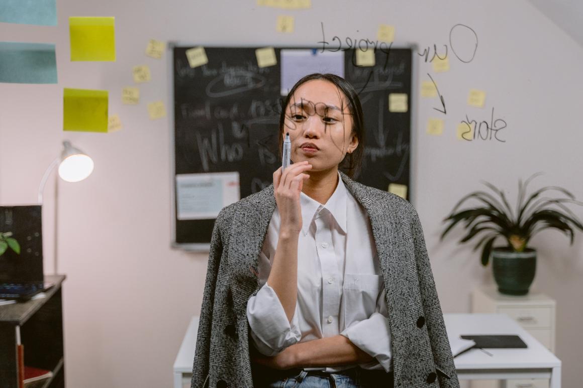 Person in a gray blazer stands thoughtfully in front of a board covered with writing and sticky notes.