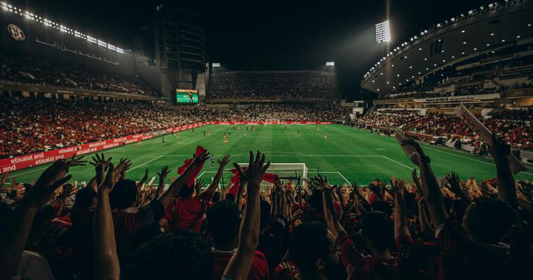 A vibrant football stadium at night, with fans in red jerseys cheering. The pitch is brightly lit, brimming with excitement and energy.