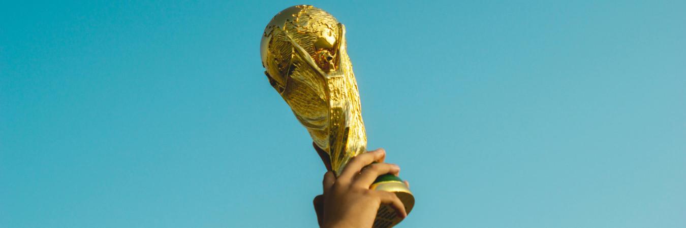 A golden trophy, resembling the FIFA World Cup, is held up against a clear blue sky.