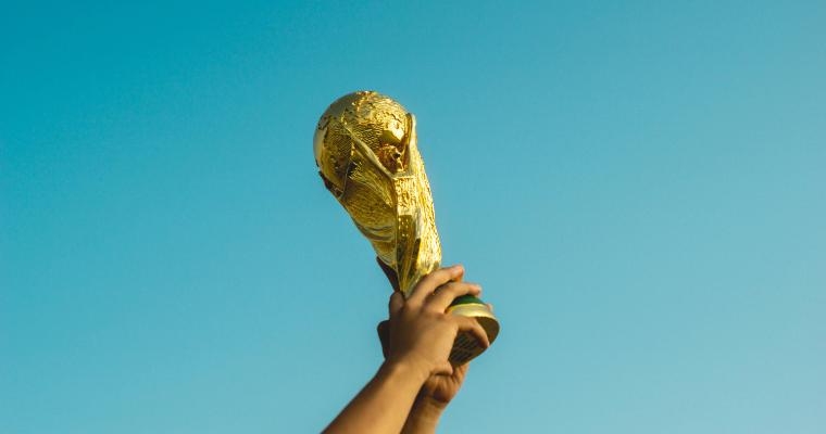 A golden trophy, resembling the FIFA World Cup, is held up against a clear blue sky.