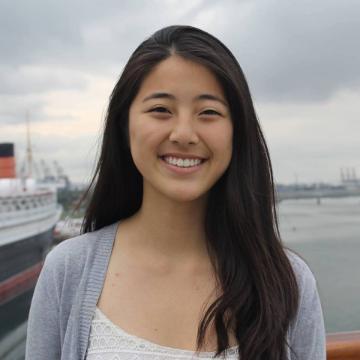 A woman smiling outdoors with a cloudy sky background.