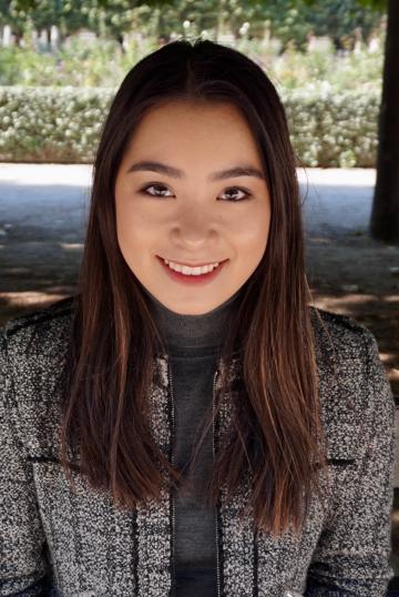 Headshot of a person smiling, wearing a grey coat and standing outdoors.