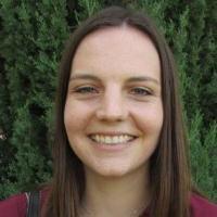 A person smiling, wearing a maroon shirt, with trees in the background.