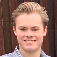 A smiling person wearing a plaid shirt in front of a wooden background.