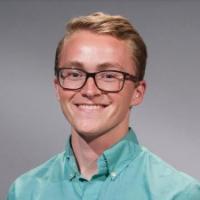 Smiling person in a teal shirt with glasses on a grey background.