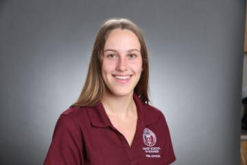 A person in a maroon shirt with an embroidered logo, smiling against a gray background.