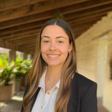 Woman in a blazer smiling outdoors near a wooden structure.