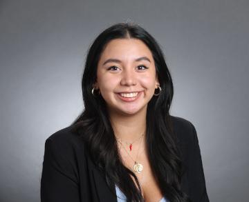 A person smiling, wearing a black blazer against a grey background.