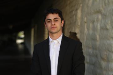 A man in a dark suit stands near a brick wall.