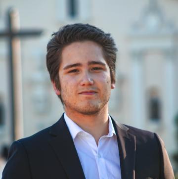 A person in a suit standing outdoors with buildings in the background.