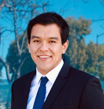 A man in a suit smiling outdoors with a blurred background.