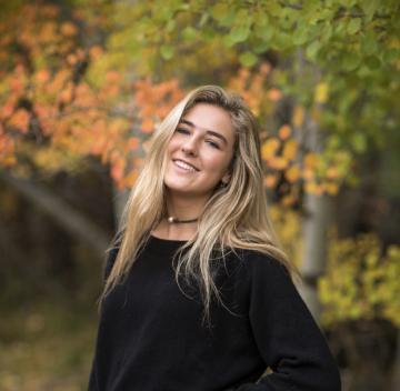 A person smiling in an outdoor setting with autumn leaves in the background.