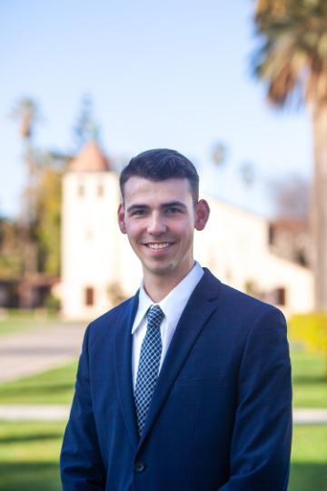 A person in a suit standing outside with buildings and trees in the background.