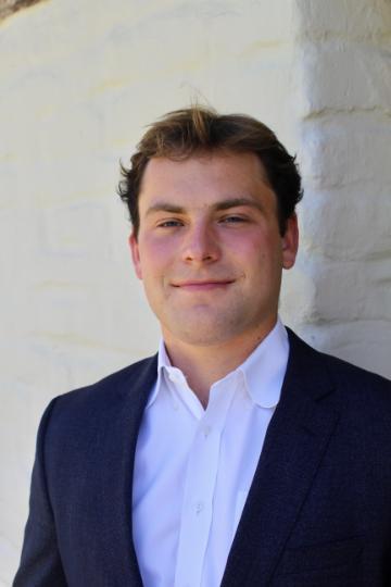 A person in a suit standing against a light-colored wall.