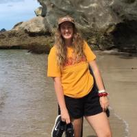 A person on a beach holding shoes with cliffs in background.