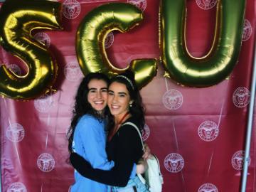 Two people hugging in front of 'SCU' balloon letters.