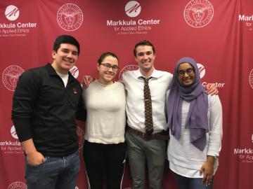Four people smiling in front of the Markkula Center for Applied Ethics backdrop.
