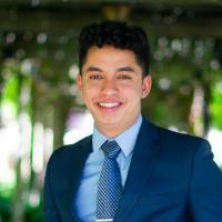 A person in a suit stands under a wooden pergola.