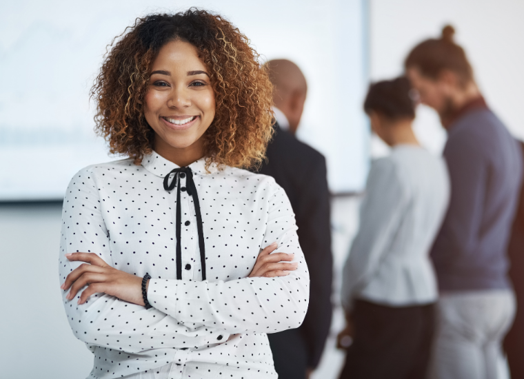 Black woman in foreground dressed in business attire with blurred colleagues standing in the background.