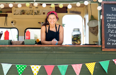 Woman entrepreneur leaning out the window of her food truck smiling. Colorful flags and lights decorate the window and part of the menu board can be seen on the right.