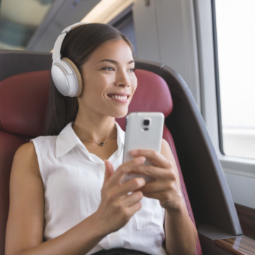 Woman riding a train with headphones, holding a smart phone and looking out the window smiling 