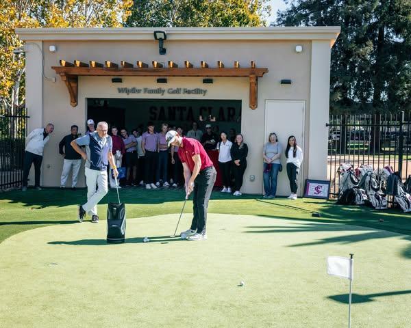 Two men take a swing on the putting green in front of a crowd at the entrance of a building with a sign that says 
