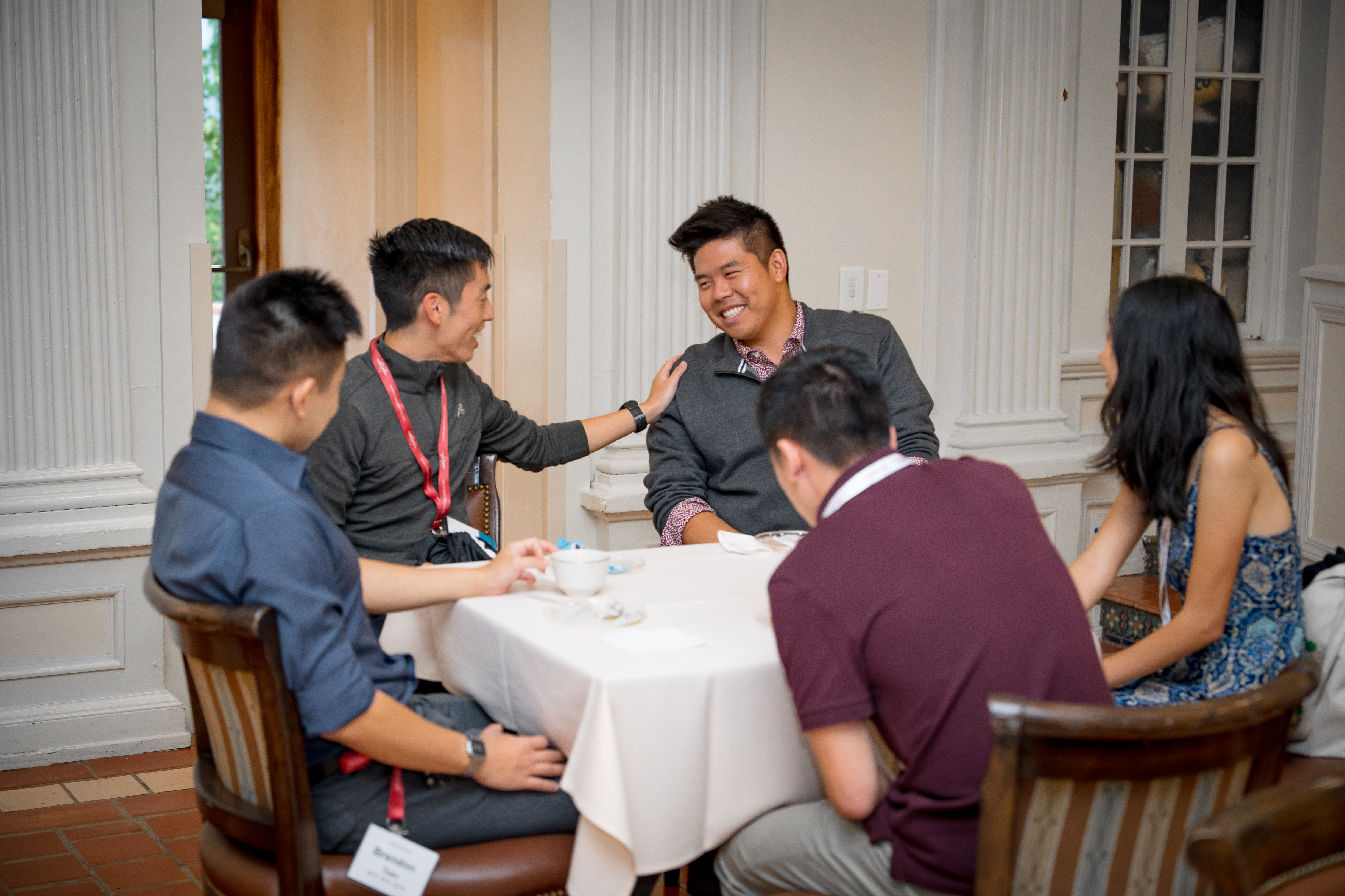 A group of five alumni seated around a table. One has his hand on the other's shoulder, who is smiling.