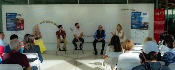 Five panelists are engaged in discussion, sitting in front of an audience