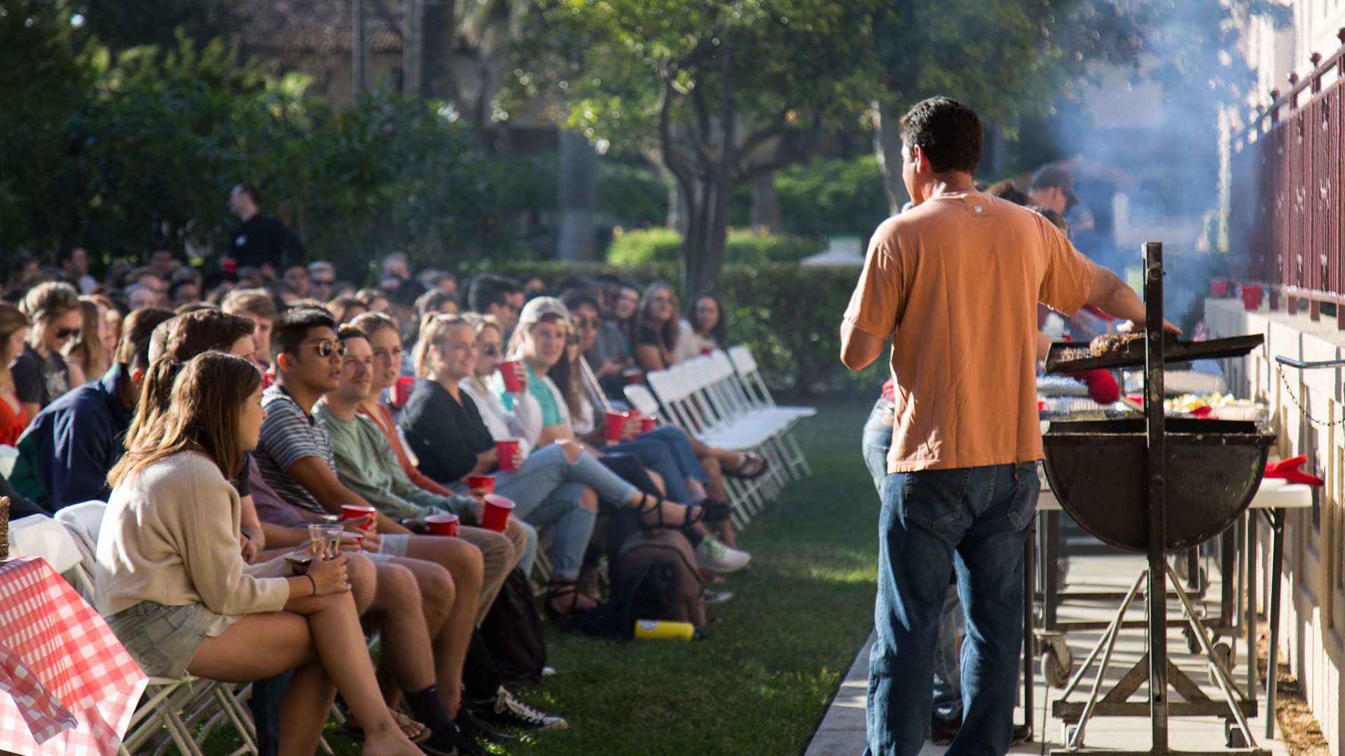 A man standing in front of a BBQ grill instructing a large group of students 