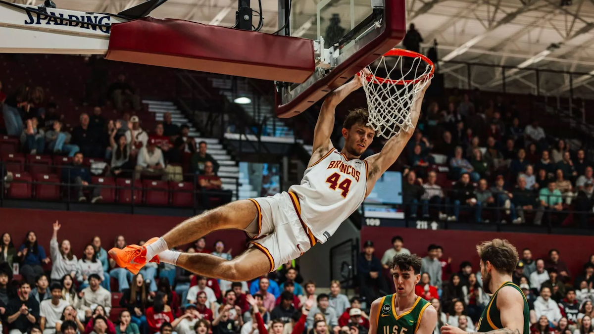 A male basketball player hangs on the rim after a slam dunk