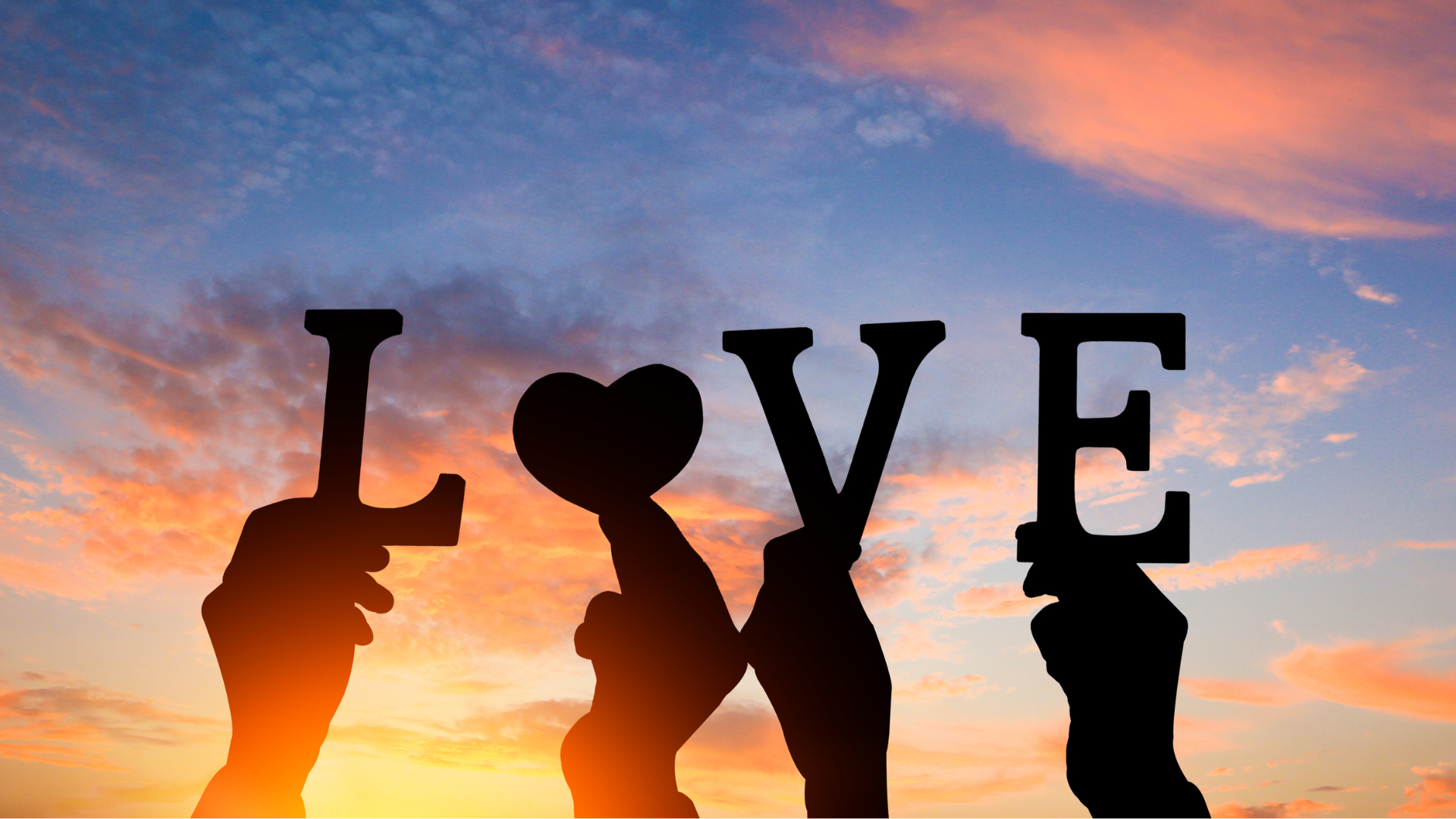 Four hands holding up letters that spell love in front of a sunset sky
