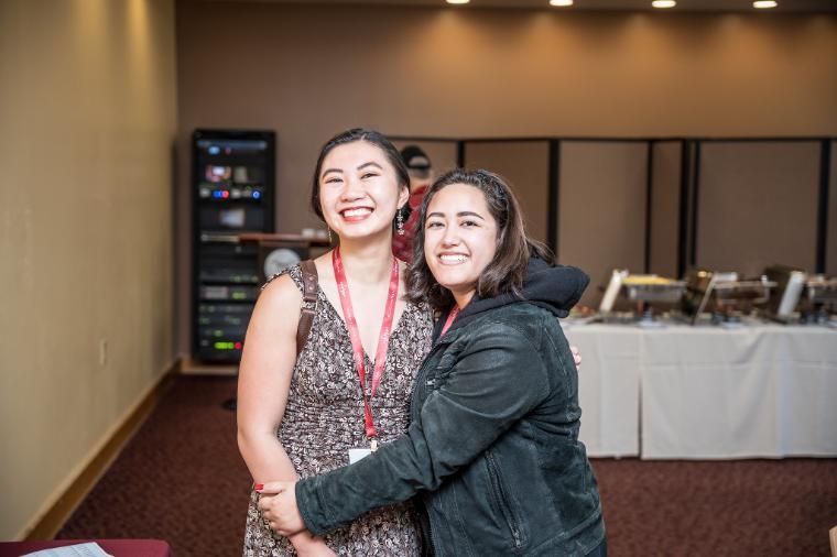 Two people hugging at an event, with tables and chairs in the background.