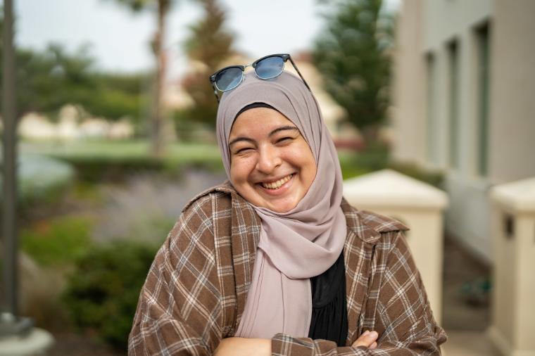 Person smiling outdoors with trees and a building in the background.