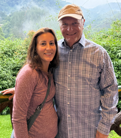 A woman and man, both smiling, standing in front of a green, leafy background.