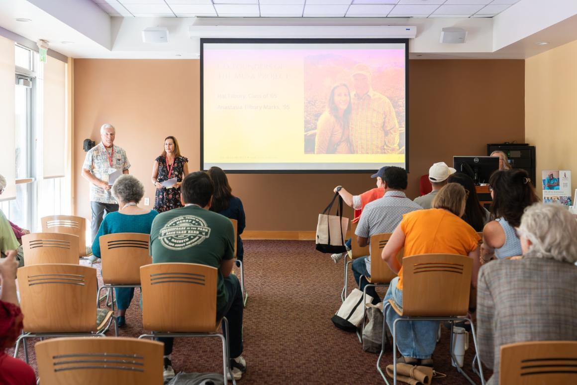 A man and a woman stand in front of a seated audience, and next to a projector screen with their photo on it, along with text reading 