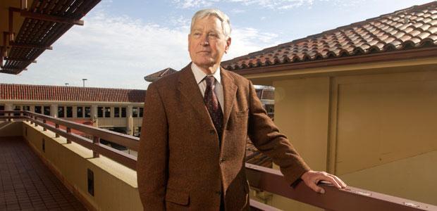 A man wearing a brown suit stands with one hand on a balcony railing, looking off to the side.