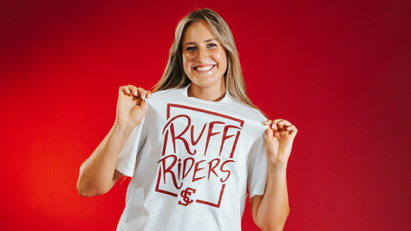 A smiling young woman against a red background. She wears a white t-shirt, holding it out from herself to show off the design, which reads 